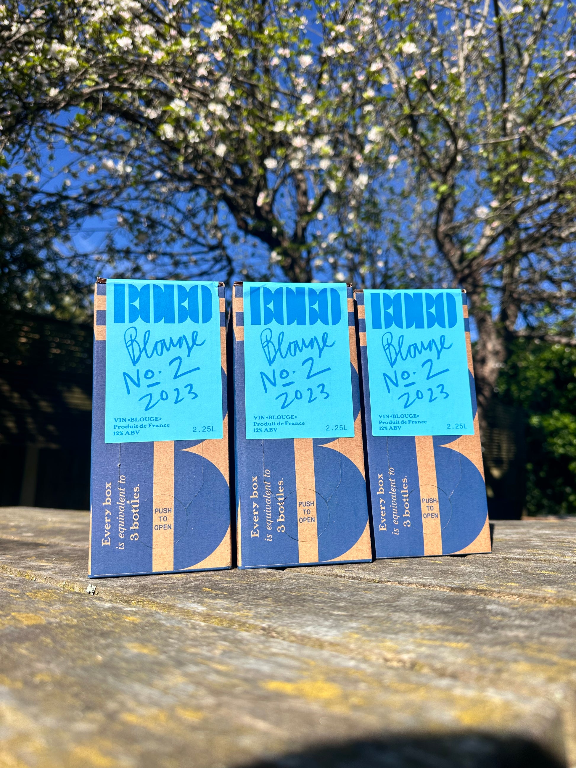 Three Bobo Blange No.2 wine boxes on a wooden table outdoors under blossom trees.